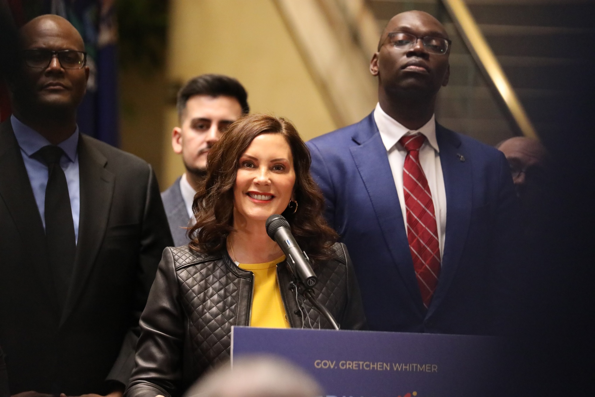 Governor Gretchen Whitmer speaking at podium with a crowd of elected officials and Michiganders standing behind them