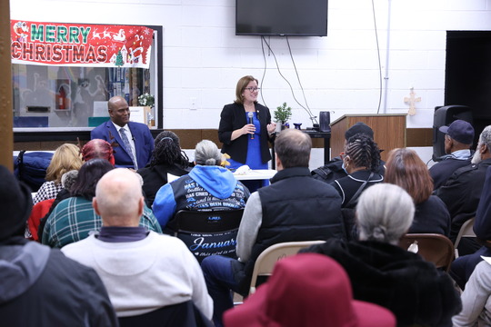 Rep. Natalie Price speaks into a microphone at the Mathis Center with Speaker Joe Tate and audience members.