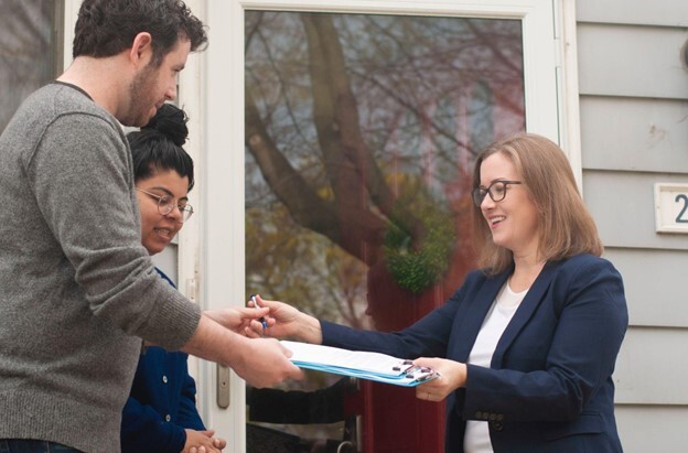 Rep. Natalie price hands clipboards to teenagers in front of a house.