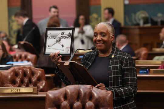 Rep. Helena Scott stands at the podium on the House Floor, holding up an black and white image in a frame of her Uncle pitching a baseball.