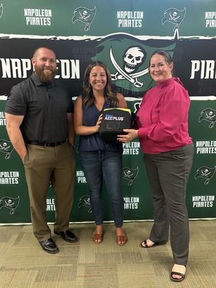 State Rep. Carrie Rheingans hands an AED to Napoleon High School officials in front of a step and repeat.