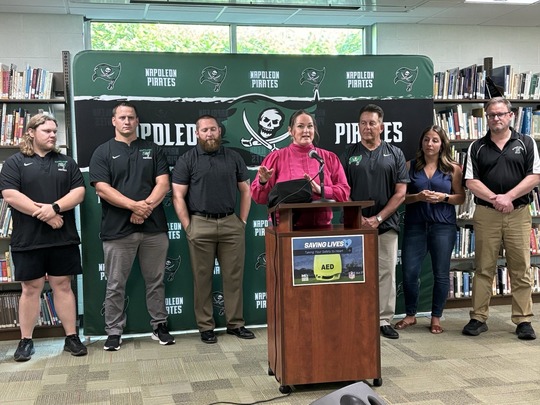State Rep. Carrie Rheingans speaks into a podium in front of Napoleon High School officials in front of a step and repeat.