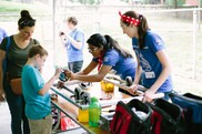 Two female volunteers leaning over table to show young boy gear, with mom standing behind him