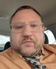 Dan sitting in his car wearing an orange shirt, a checkered tie, and clear-framed glasses