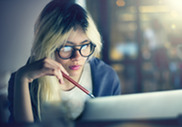 Blonde woman in glasses holding pencil in one hand while looking at laptop screen