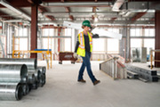 Woman in a hardhat and yellow vest carrying a board while walking through a room under construction