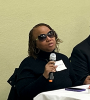 Shawnese Laury wearing a black sweater and sunglasses while holding a microphone and speaking