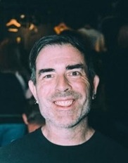Chris Roach smiling while wearing a black sweater against a dark background