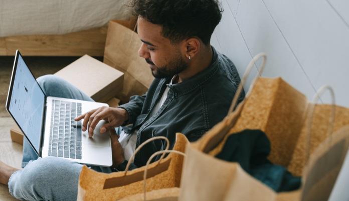 man online shopping seated with two brown bags with handles next to him