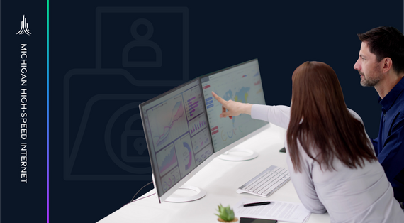 woman and man sit at desk analyzing data on two monitors while woman points at one monitor