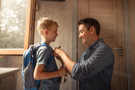 A father helps his son with his schoolbag at the front door.