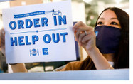 Photo of a restaurant worker hanging up a sign reading "Order In / Help Out"