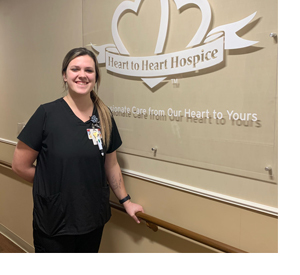 Gracie Gabalis, a healthcare professional standing in front of a wall sign reading Heart to Heart Hospice: Compassionate Care from Our Heart to Yours