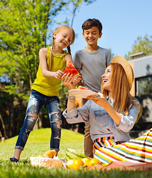 Woman in a colorful summer skirt sharing a plate of watermelon with two school age children