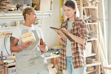 Two people standing in a woodshop one with a notebook and the other holding a box