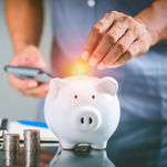 a man's hand putting a coin in a piggy bank, with stacks of coins nearby