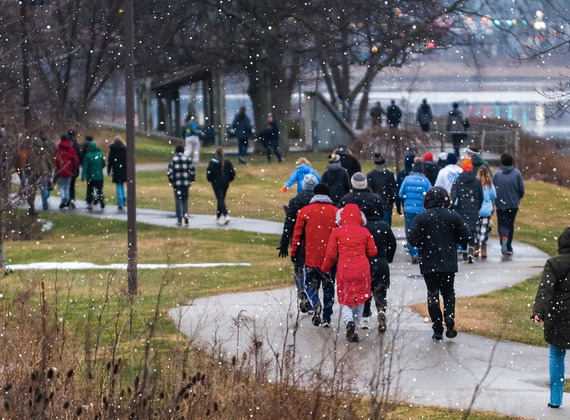 Snow falls as a Walk for Warmth group in winter coats walks along a winding paved path through a park.