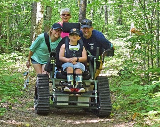 A boy in an All-Terrain Track Chair poses with family members while on the trail