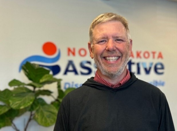Mike Chaussee, a white man with blonde hair smiles in front of the North Dakota Assistive logo