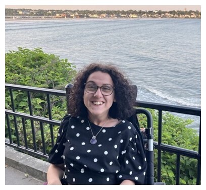 A woman with curly brown hair and glasses is smiling. She is seated in a wheelchair. She has a black top with white polka dots and a pendant necklace