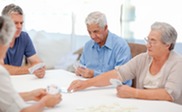 Group of four older adults playing cards at table, with windows behind them. 