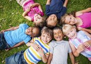 7 young, smiling children wearing colorful shirts laying on green grass in a circle, with their heads in the middle. 
