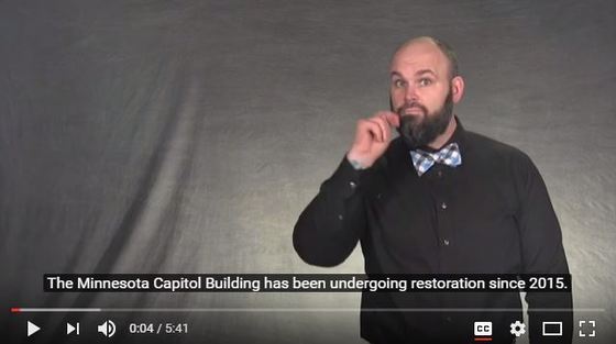 Still frame from the accessibility at the MN Capitol video. Justin Small is signing, "Minnesota." He is wearing a black shirt and a plaid bow tie.