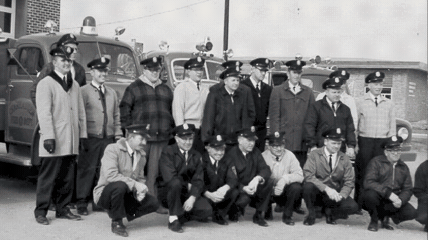Historic photo of Chanhassen firefighters transitions into a modern fire truck leaving the station as a firefighter waves and smiles at the camera.