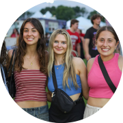 Five young women smile for a photo at an outdoor community festival with carnival booths and crowds in the background.