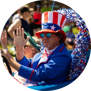 Person dressed as Uncle Sam waves from a blue parade car as spectators watch along the route.