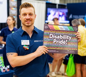 Trevor Turner is wearing a blue shirt and smiling while holding a Disability Pride poster.