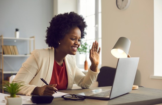 Smiling woman sitting at desk waving at computer screen