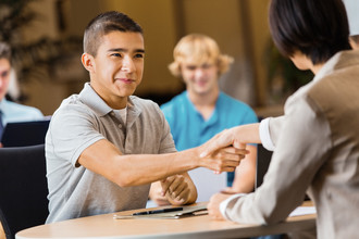 Two teen boys sitting across a desk from each other, shaking hands.