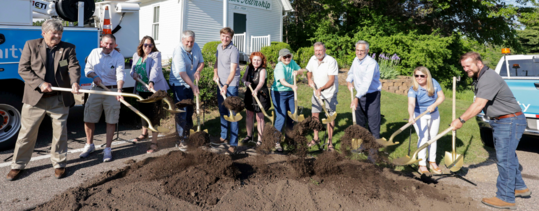 A group of ceremony attendees stand near broken dirt, using shovels, in Stillwater Township
