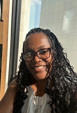 A woman with long curly hair, wearing glasses and a sleeveless top with stripes, smiles while sitting by a window.