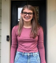 Ava smiling, wearing glasses, a red long-sleeve shirt and blue jeans with her hair down.