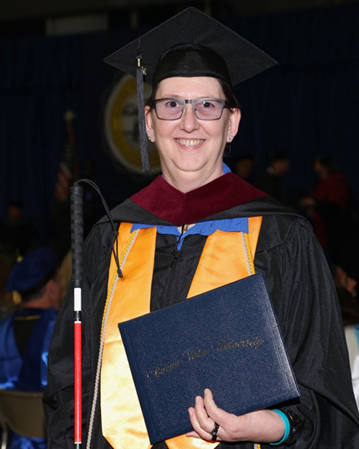 Laurie smiling, dressed in a black graduation cap and gown. Laurie is holding a diploma in one hand and a cane in the other.