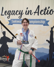 with dark brown hair, wearing black glasses, She is wearing a Taekwondo Gi (uniform) with a green belt.