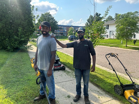 Two men stand surrounded by lawn care equipment, smiling for a picture.