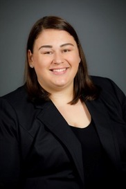 Professional portrait of Lindsay smiling with shoulder-length brown hair wearing a black blazer against a dark gray background