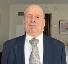 Steve looking at the camera wearing a navy suit, white dress shirt and blue and grey patterned tie standing indoors. 