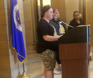 Teenage boy in T-shirt and shorts speaks at podium near Minnesota flag. Two other teens stand beside him.