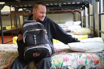 man with backpack sitting on bed in homeless shelter