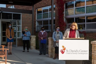 Commissioner Harpstead speaking at a podium behind a sign saying St. David's Center, while people in the background watch.