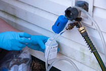 Gloved hands sampling water through a filter at a hose spigot