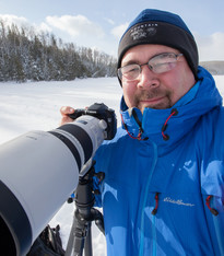 Man smiling and looking at the camera holding a camera with a long lens with pines and snow in the background