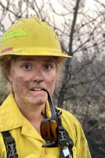Female firefighter with hard hat and yellow jacket, looking at camera, holding sunglasses in her mouth.