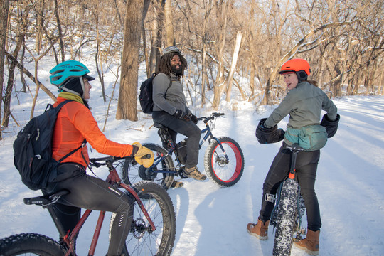 A Latino woman, a Black man, and a white woman on their fat bikes on a snowy trail.