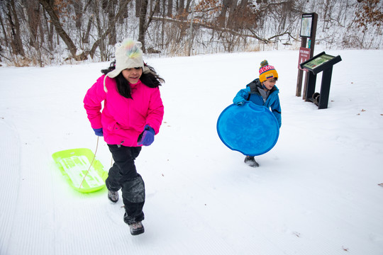Latino girl and her little brother smiling while pulling their sleds up a snowy hill.