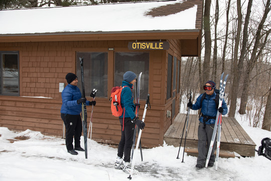 A Black woman, an Southeast Asian woman and a Filipino man holding skis by camper cabin in the woods.